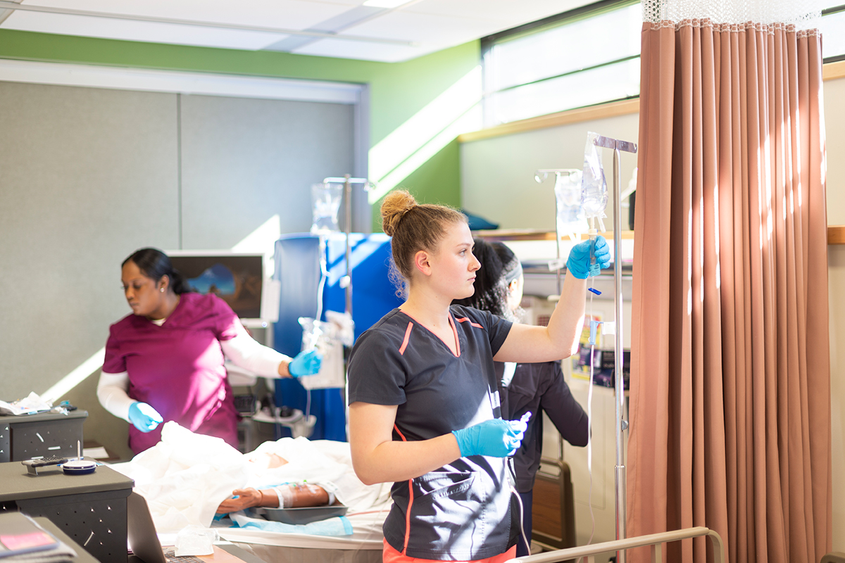 A student examines an IV unit, while other students assist a patient..