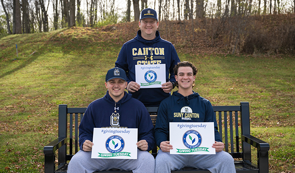 Three baseball players holding Giving Tuesday signs sit on a bench outside French Hall.