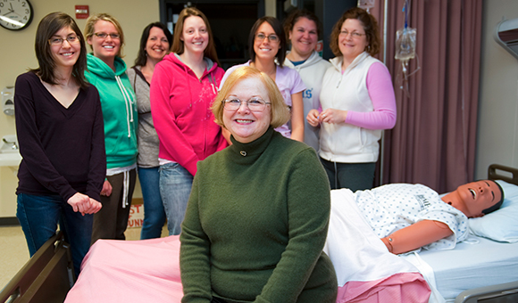 Linda Fay is surrounded by Nursing students as she sits on a patient bed in the Nursing lab.