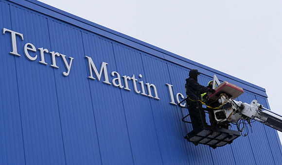 Workers install the Terry Martin Arena signage on the blue exterior of Roos House.