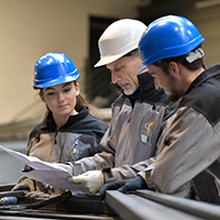 Two students with blue hard hats review plans with a professor.