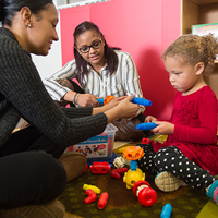 Two daycare professionals assist a toddler with connection toys.