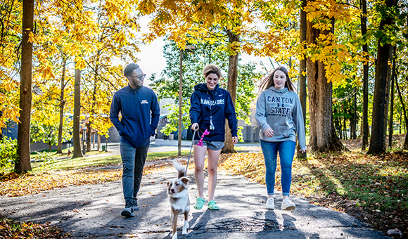 Three students walking a dog from the residence halls on a fall day.