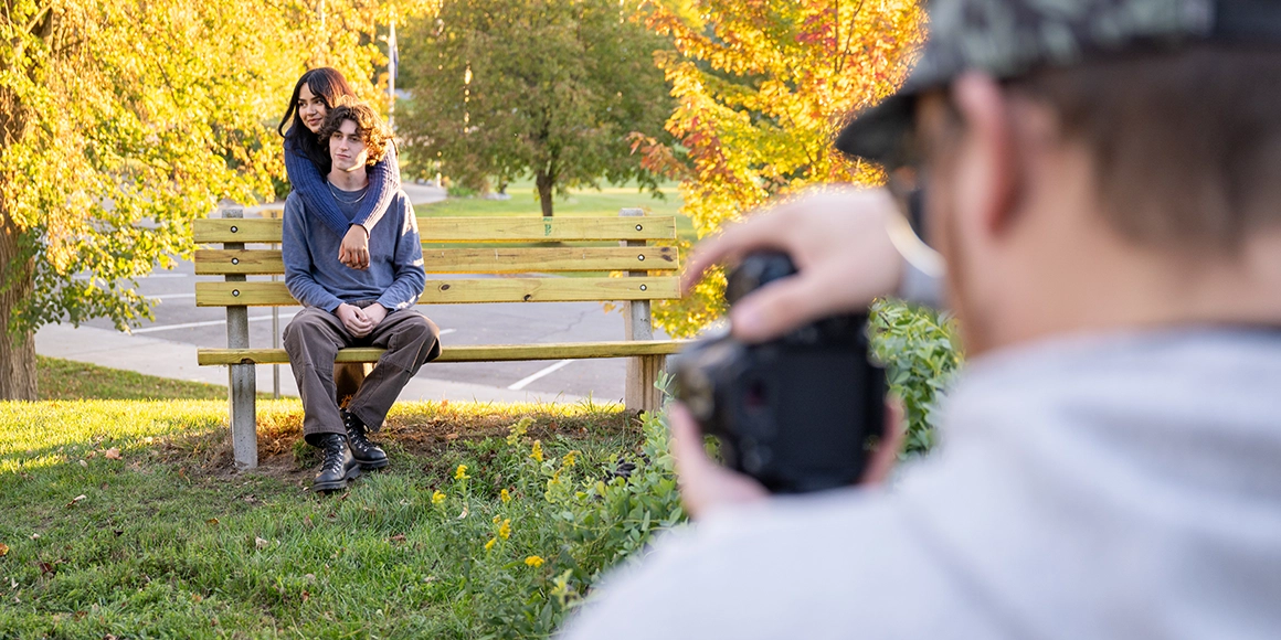 Two students pose on a bench during the golden hour while another photographs them.