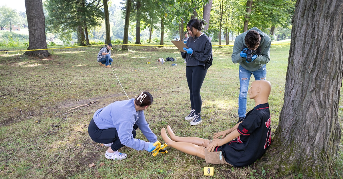 A student photographs a dummy victim while others measure and take notes of a mock crime scene.