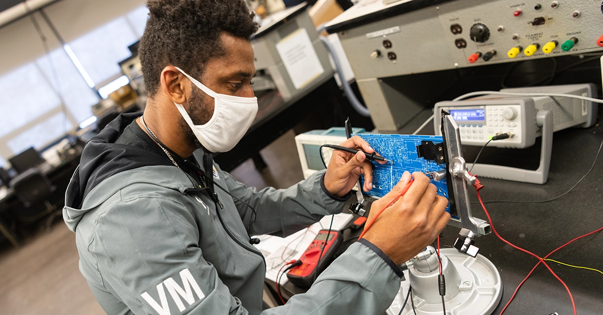 A student wearing a mask solders a motherboard in the Electrical Technology lab.