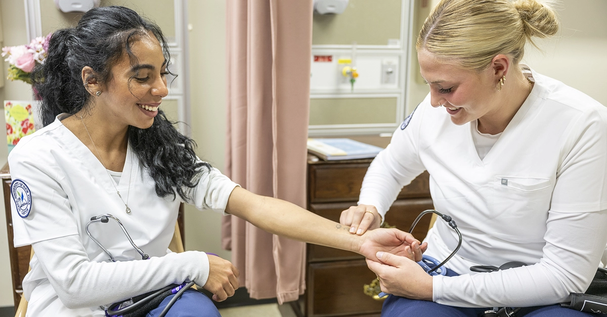 A nursing student checks the pulse on another student in a nursing lab.
