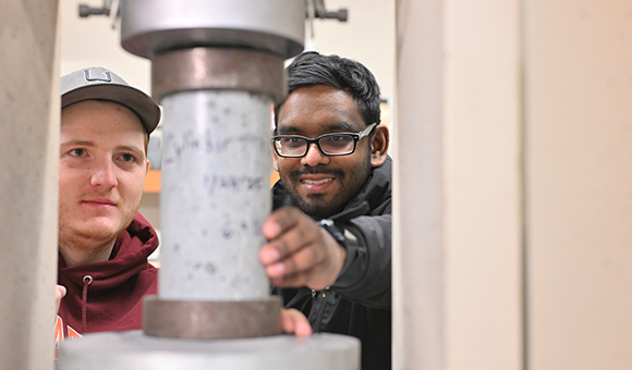 Mathew Greene and Chandradat Rampat place a lightweight concrete cylinder in a hydraulic press for testing.