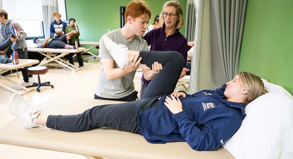 A student stretches a patient in the Physical Therapist lab while Instructional Support Associate Anne Reilly looks on.