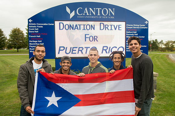 Five students hold a Puerto Rican flag in front of the SUNY Canton digital sign.