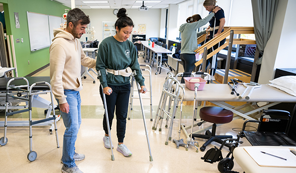 A student assists another student with assistive crutches while other students work on stretching in the background.
