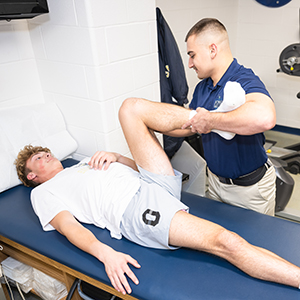 Physical Therapist Assistant intern Connor Dickinson helps to stretch a student athlete's leg on a table.