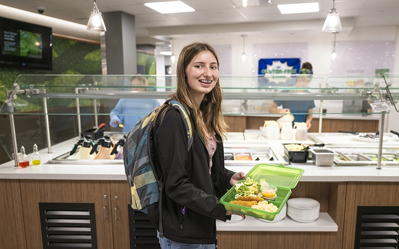 A student holds up an OZZI box filled with salad, chicken tenders and macaroni and cheese.