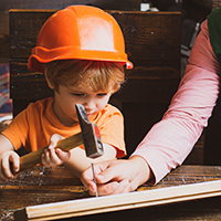 A young student wearing an orange hard hat hammers a nail with the help of an adult.