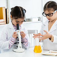 A young camper looks through a microscope while another examines a chemistry vial with yellow liquid.